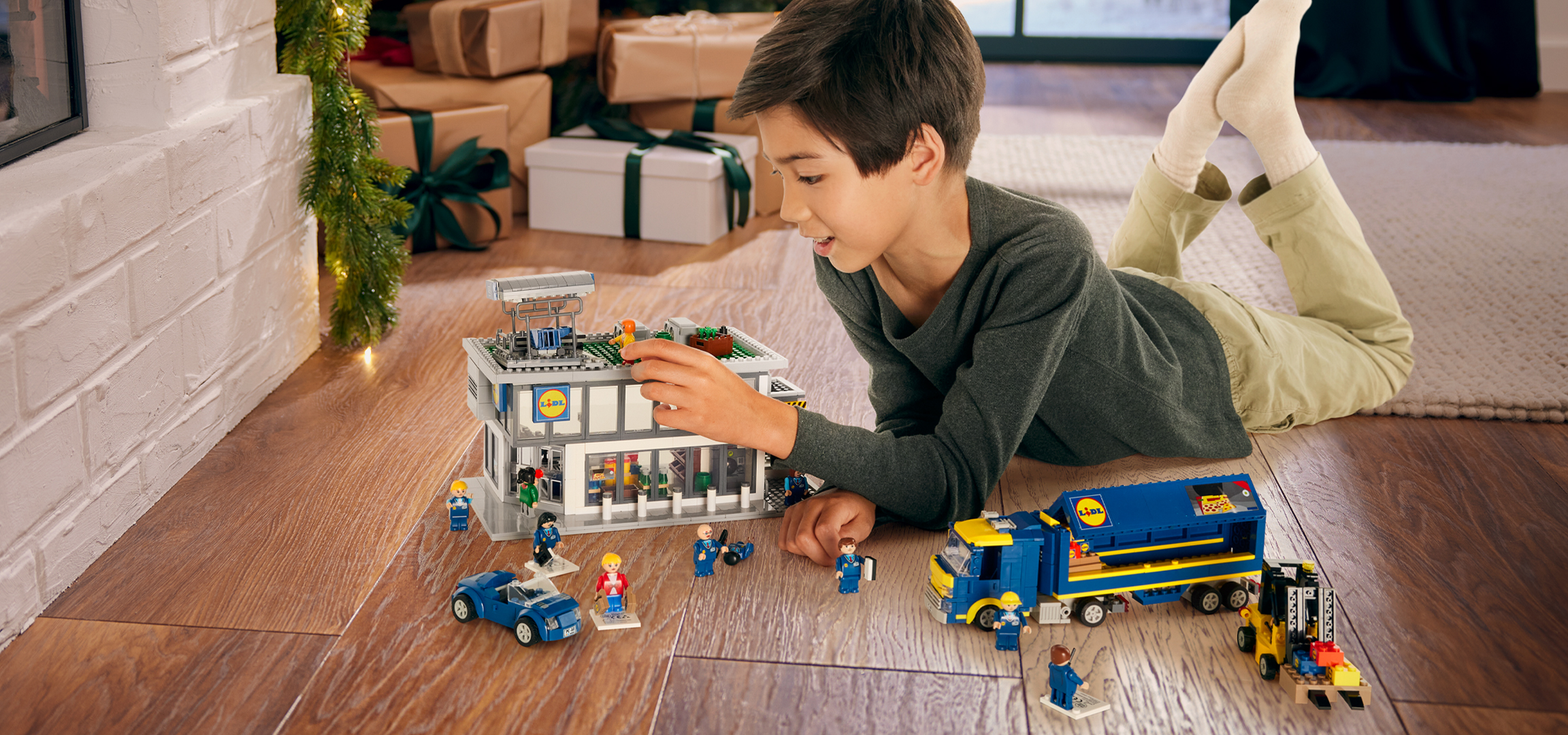 Fiú játszik Lidl LEGO készlettel, beleértve egy boltot, teherautót és figurákat, karácsonyi dekorációk mellett.