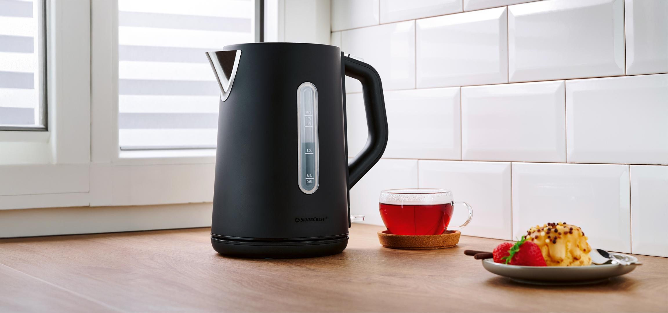 Fekete SilverCrest vízforraló, teáscsésze és sütemény egy konyhapulton.