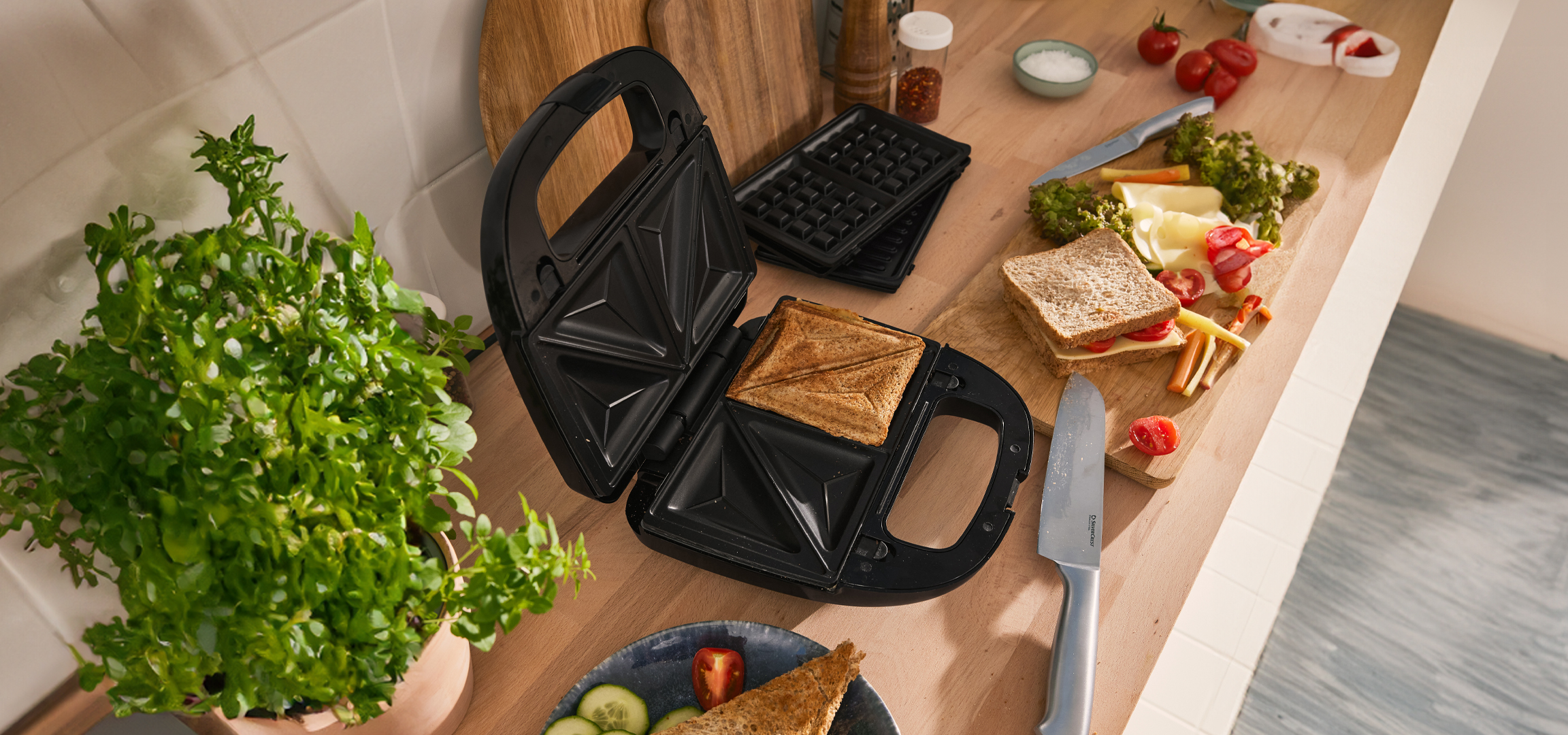 Szendvicssütő, gofrisütő és grillező lapok, friss szendvics hozzávalók egy fa vágódeszkán.