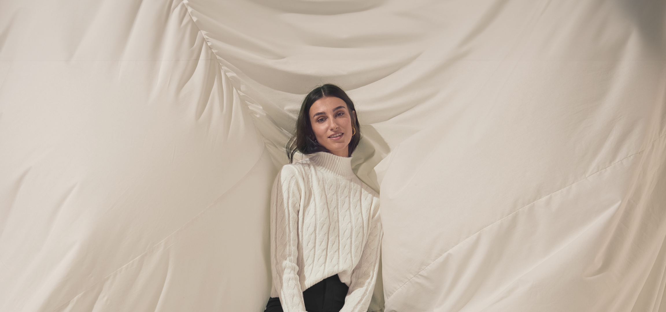 Nő fehér pulóverben és fekete nadrágban, világos háttér előtt.