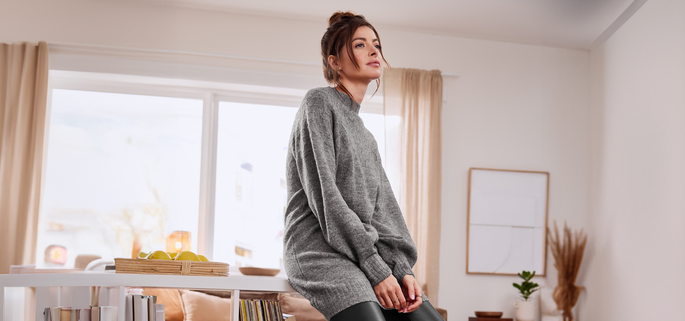 Nő szürke pulóverben, fehér polcokkal és ablakokkal teli szobában.