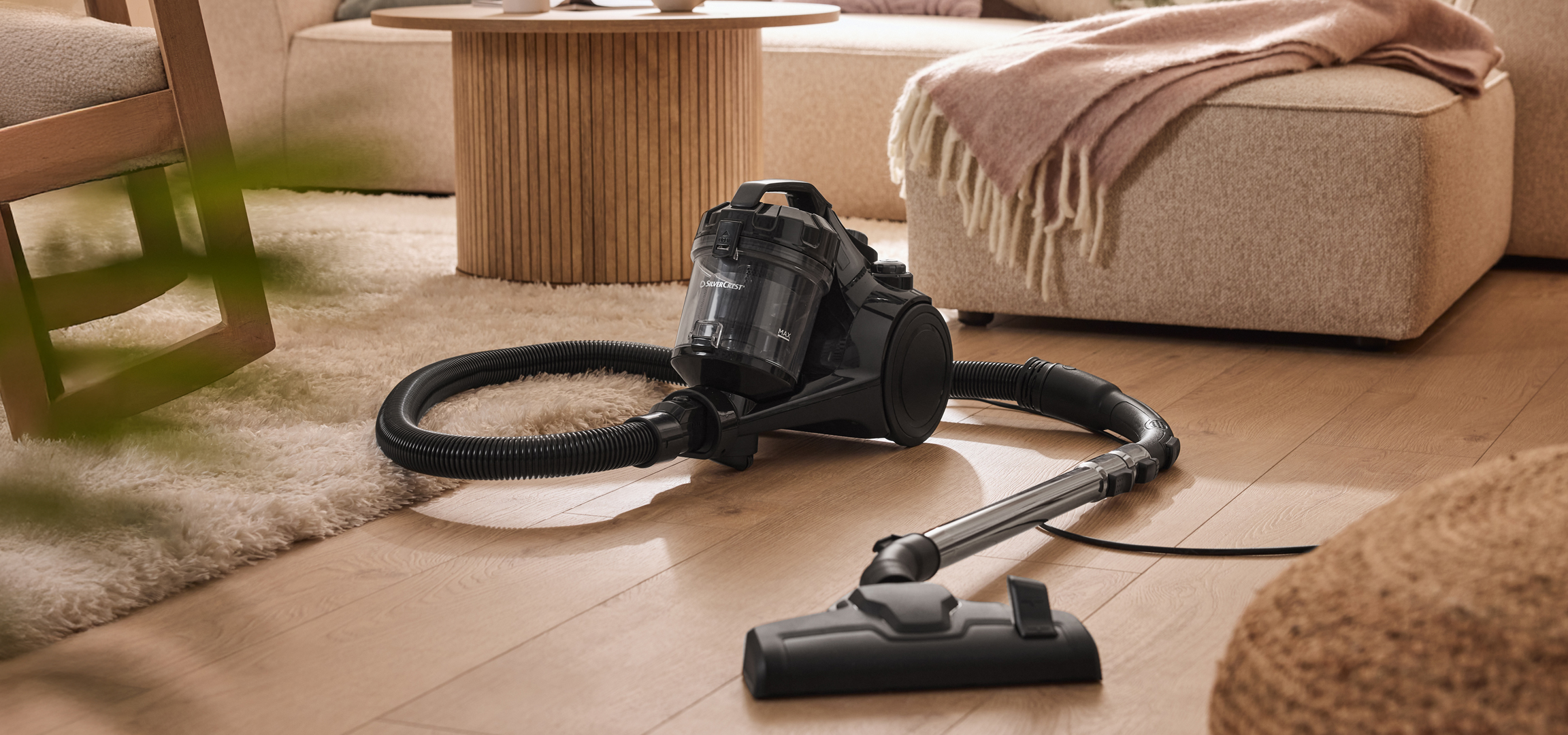 Fekete SilverCrest porszívó egy világos szőnyegen és fapadlón, növényekkel a háttérben.