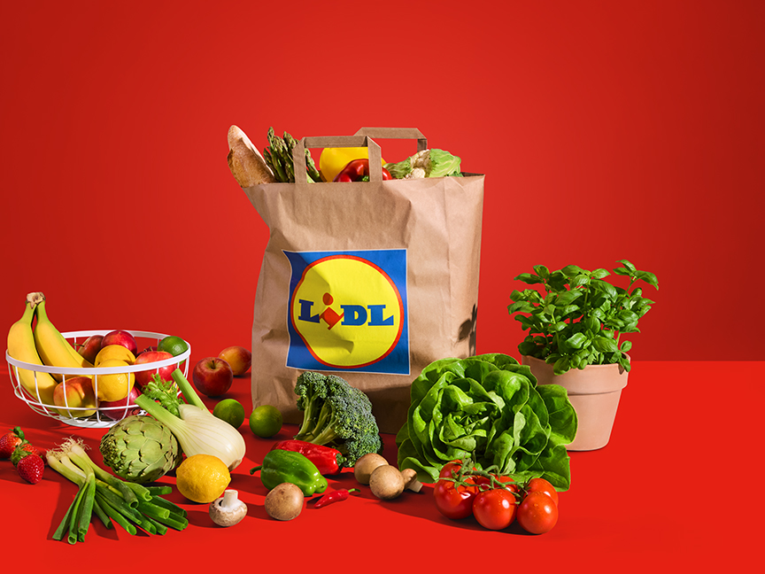 Lidl bevásárlószatyrot telepakoltak friss zöldségekkel és gyümölcsökkel egy piros háttér előtt.