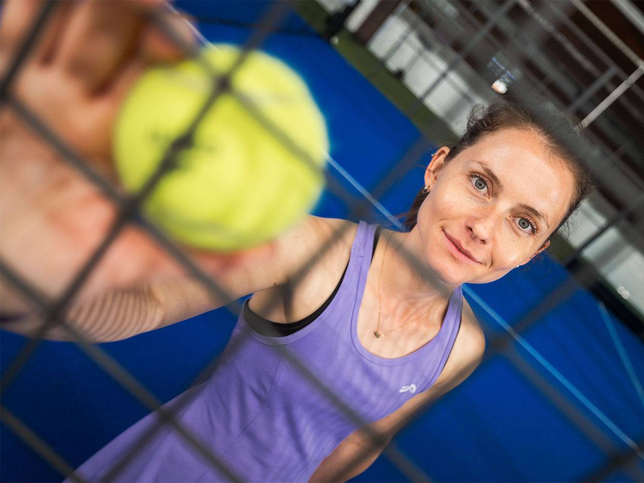 Nő sportruházatban padellabdát tart a hálón keresztül, kék pálya a háttérben.