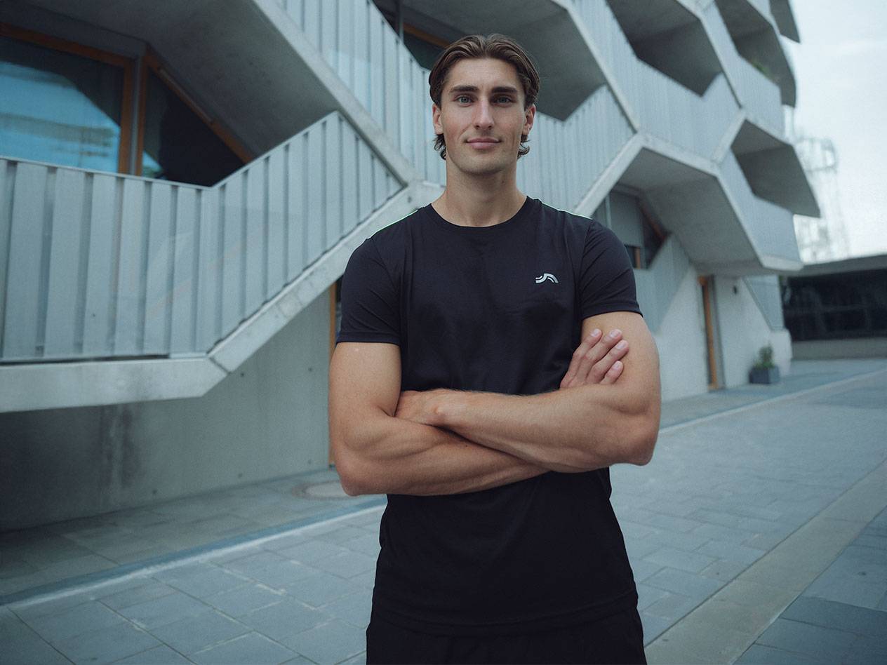 Férfi fekete logós pólóban és fekete rövidnadrágban, karba tett kézzel áll a szabadban.