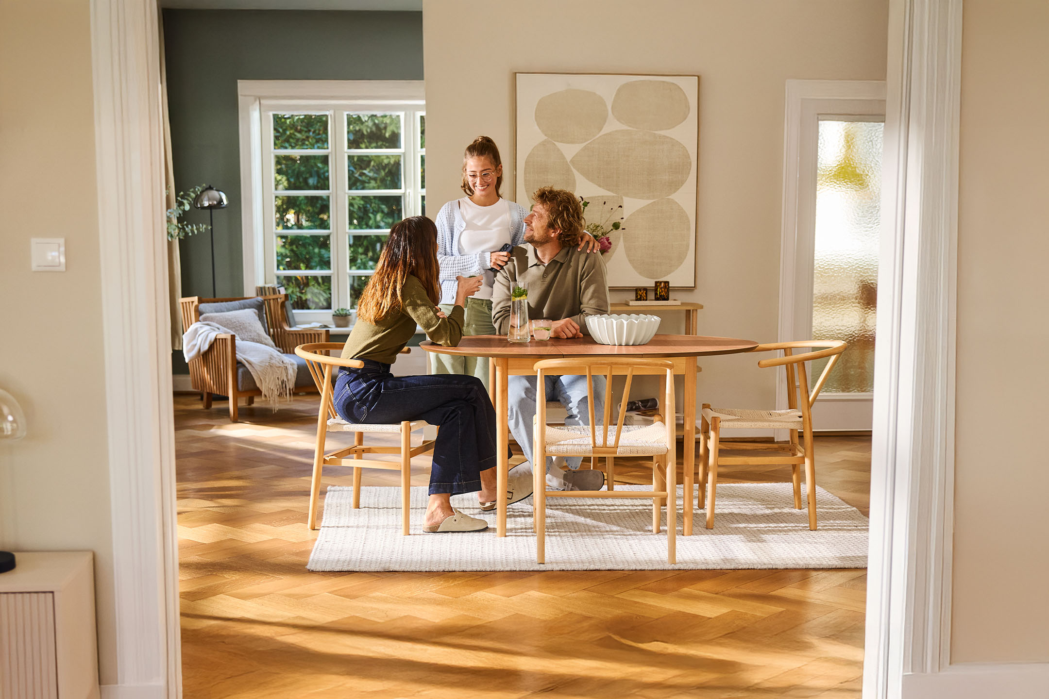 Három ember beszélget egy fa asztalnál egy világos nappaliban, nagy ablakokkal.