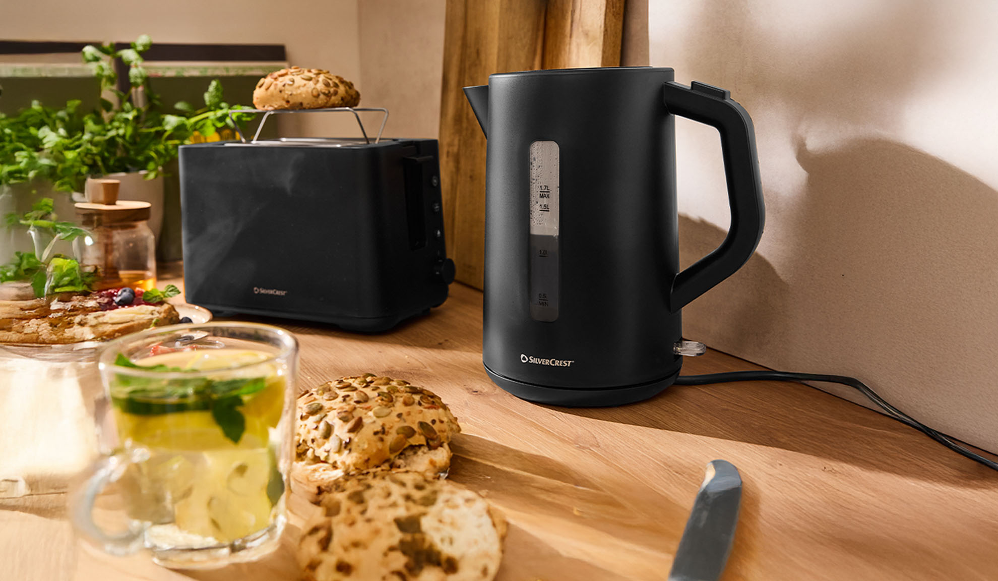 Fekete SilverCrest vízforraló és kenyérpirító fa asztalon reggelivel.