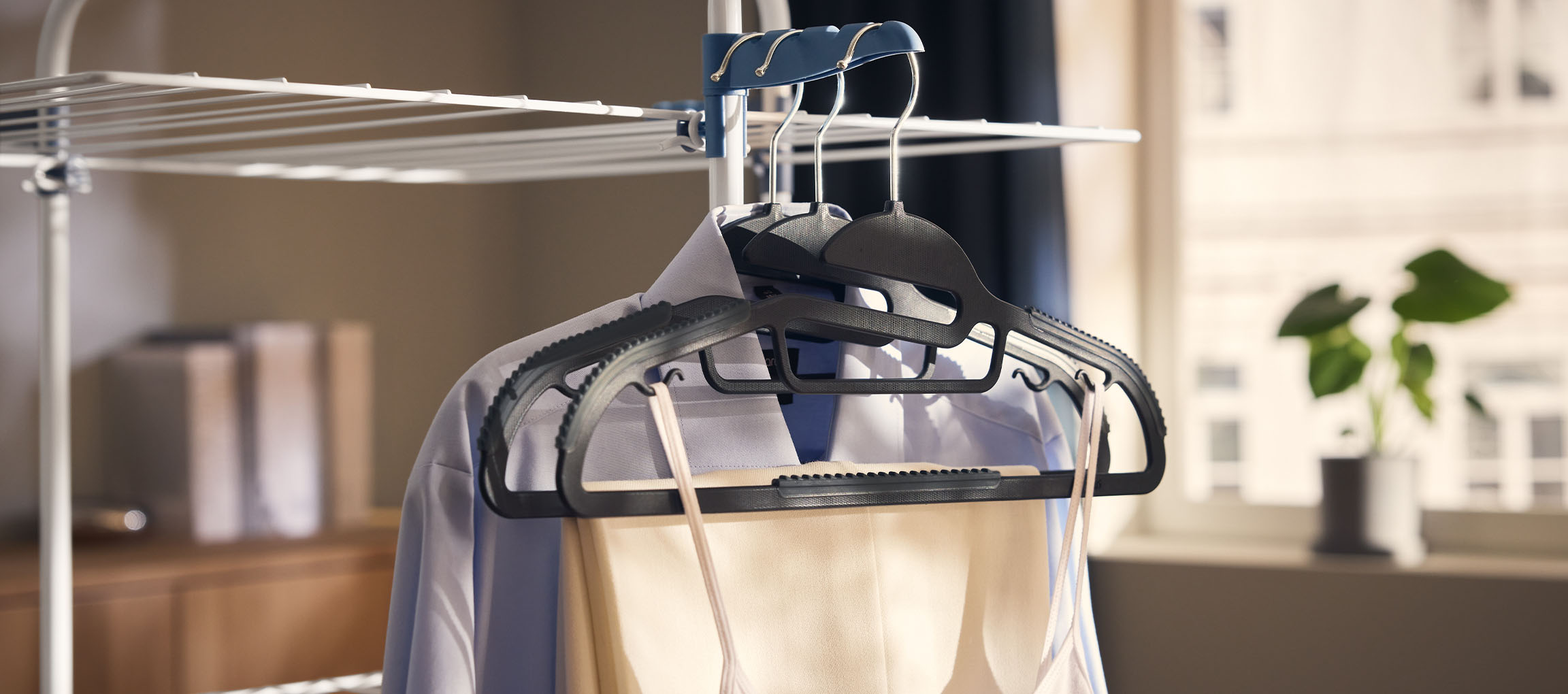 Ruhák vállfákon egy szárítóállványon, növénnyel a háttérben.