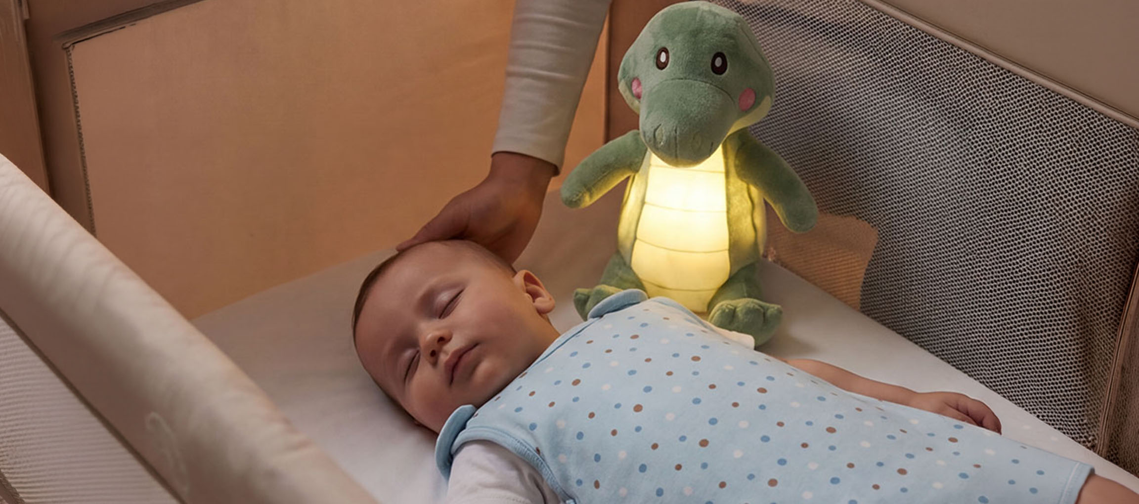 Baba alszik kiságyban plüss éjszakai lámpával, felnőtt simogatja a baba fejét.