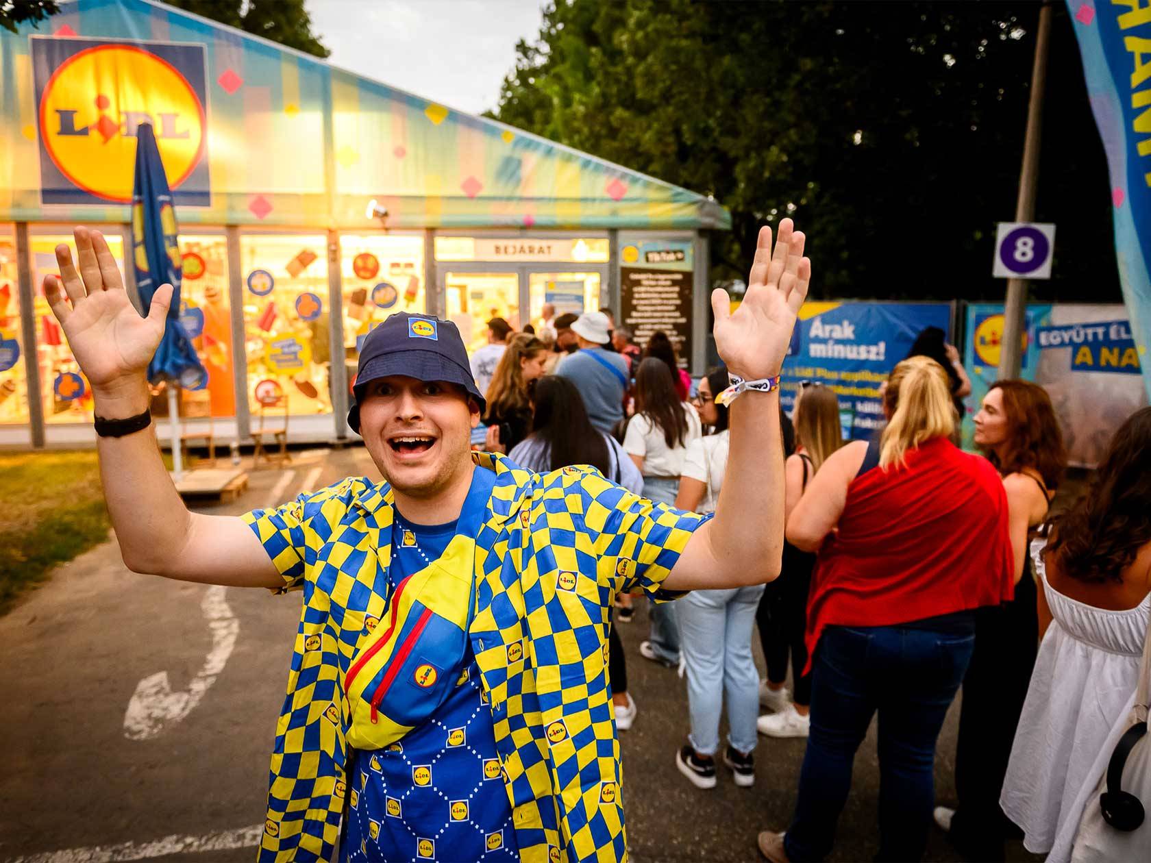 Lidl-es pólóban és sapkában lévő férfi integet a Lidl fesztiválsátor előtt.