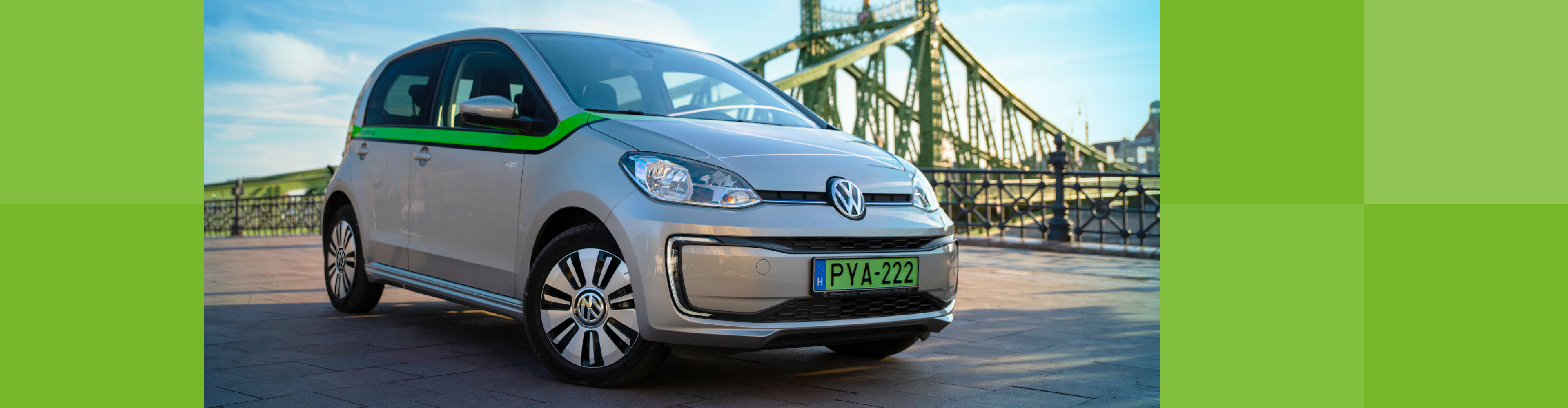 Ezüstszínű Volkswagen e-Up autó zöld csíkkal, háttérben a Szabadság híddal.