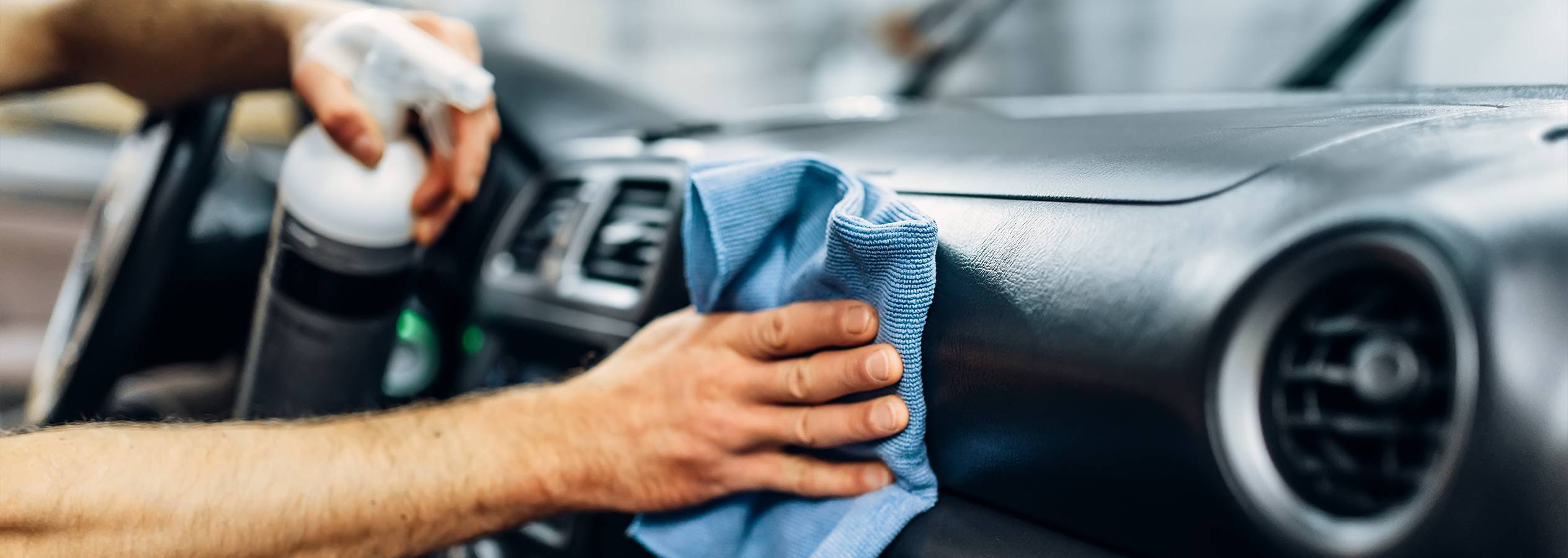 Egy személy tisztítja az autó műszerfalát egy spray-palackkal és egy kék mikroszálas kendővel.