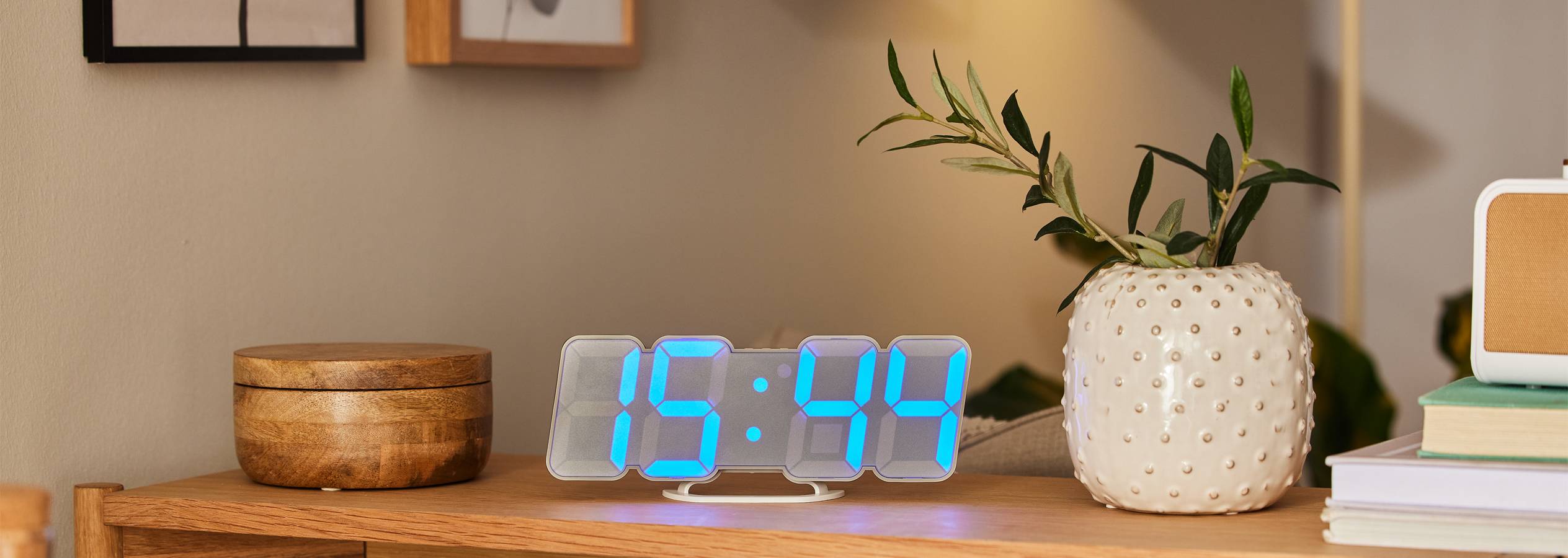 Modern LED digitális óra egy fa asztalon, virágcseréppel és könyvekkel a háttérben.