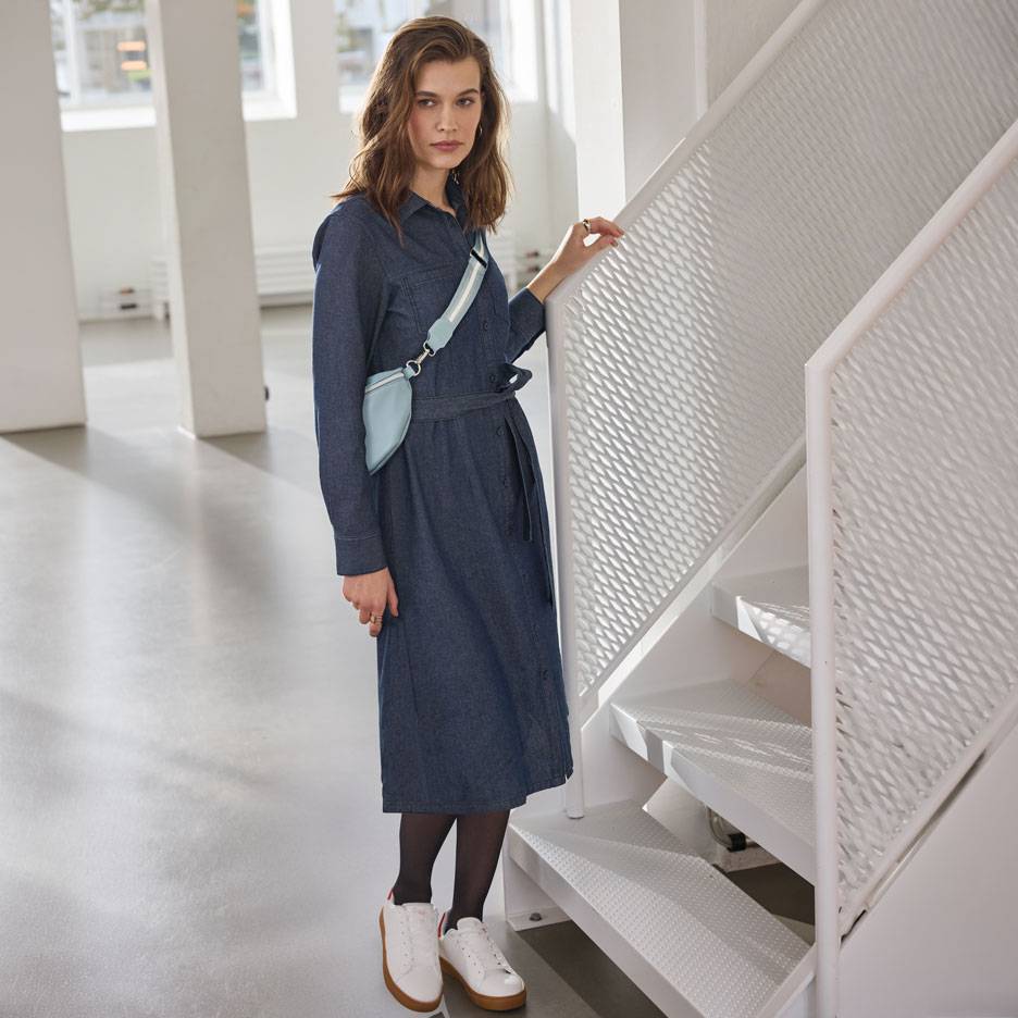 Nő farmer ingruhában, fekete harisnyában és fehér sportcipőben, világoskék övtáskával.