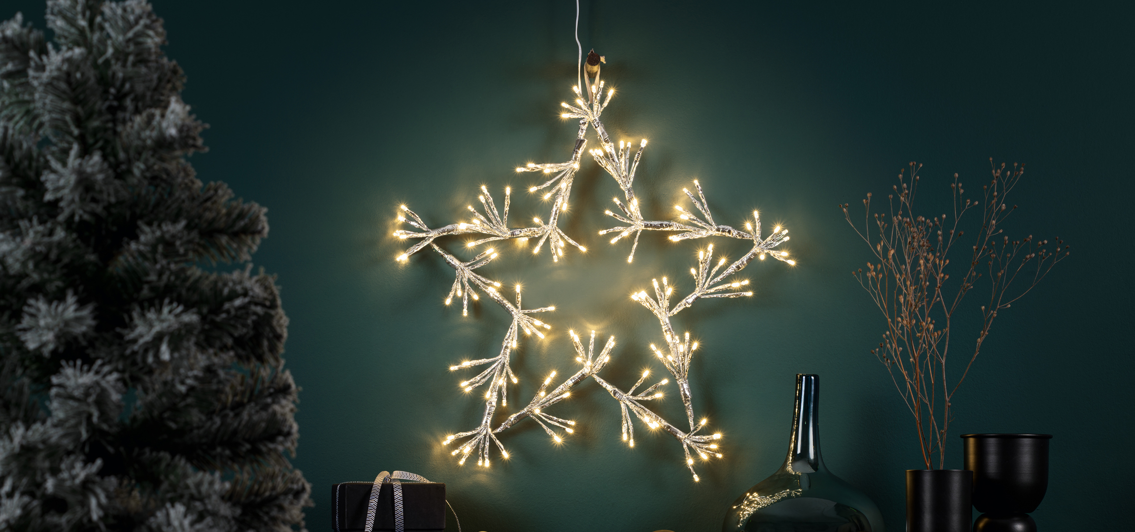 Karácsonyi dekoráció műfenyővel, világító csillaggal és vázákkal sötétzöld háttér előtt.